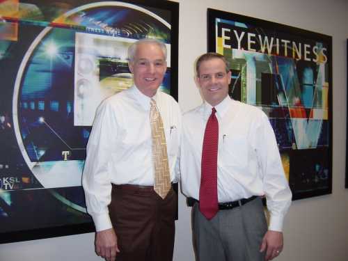 Two men in dress shirts and ties stand smiling in front of colorful television station graphics.