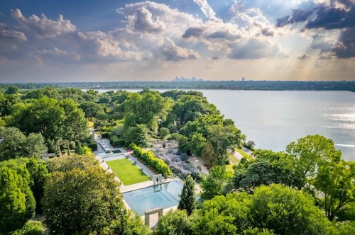 Aerial view of a lush landscape with a pool, gardens, and a lake, under a cloudy sky with sunlight breaking through.
