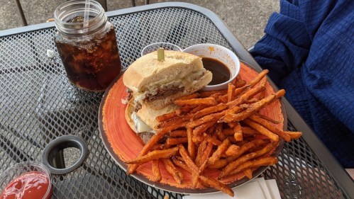 A plate with a sandwich and a side of sweet potato fries, accompanied by a drink in a jar and dipping sauce.