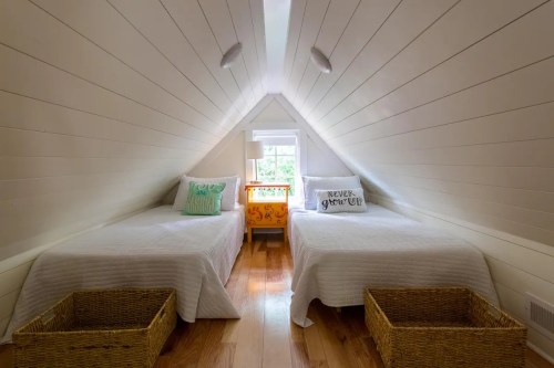 Cozy attic bedroom with two twin beds, white walls, and woven baskets, featuring a bright window and decorative pillows.