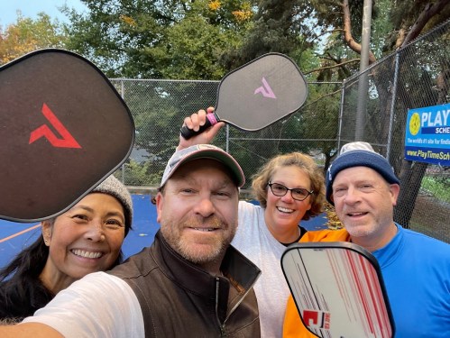 A group of four people smiling and holding pickleball paddles on a court, surrounded by trees and a fence.