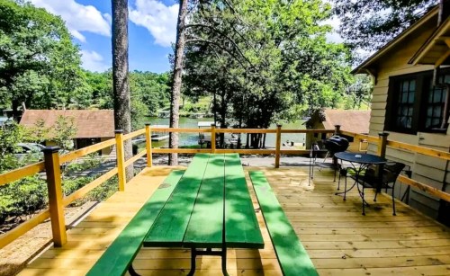 A green picnic table on a wooden deck overlooking a serene lake surrounded by trees and cottages.
