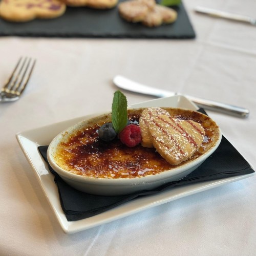 A creamy crème brûlée topped with a mint leaf, served with a cookie and fresh berries on a white plate.