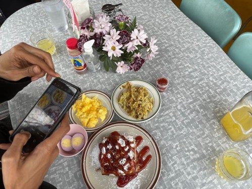 A table set with a floral centerpiece, a plate of waffles with syrup, scrambled eggs, and a person taking a photo with a phone.