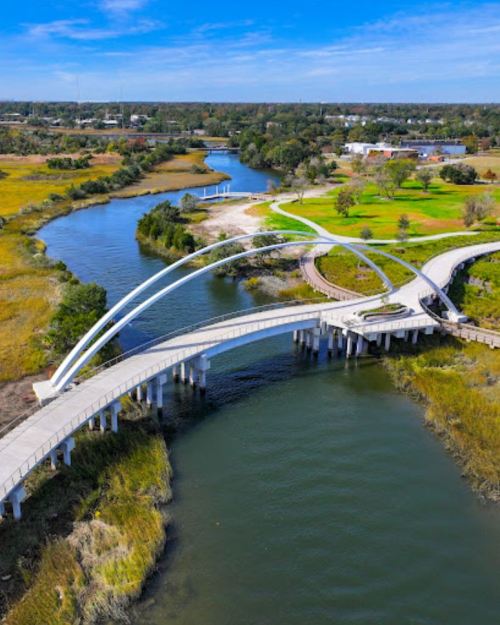 Aerial view of a modern bridge over a river, surrounded by green parks and a clear blue sky.