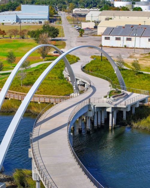 A modern curved pedestrian bridge spans a waterway, surrounded by green grass and industrial buildings in the background.