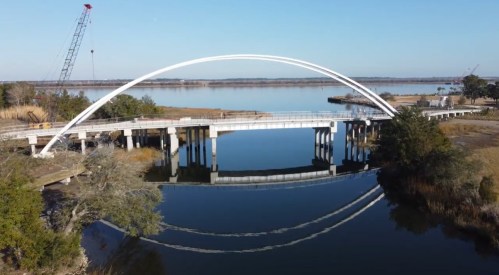 A modern white arch bridge spans a calm river, reflecting in the water, with cranes and trees in the background.