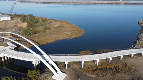 Aerial view of a curved bridge over calm water, surrounded by grassy banks and trees.