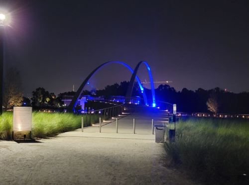 A lit blue arch bridge at night, surrounded by greenery and a pathway leading towards it.
