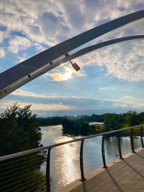 A scenic view of a river under a cloudy sky, with a modern bridge arching overhead. Sunlight reflects on the water.
