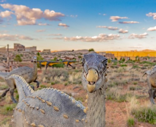 A close-up of a dinosaur sculpture in a desert landscape, with a colorful sky and distant structures in the background.