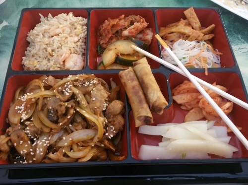 A colorful bento box featuring fried rice, kimchi, spring rolls, and various side dishes.