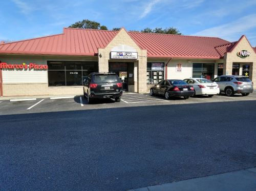 A strip mall with several businesses, including Marco's Pizza, and parked cars in front on a sunny day.