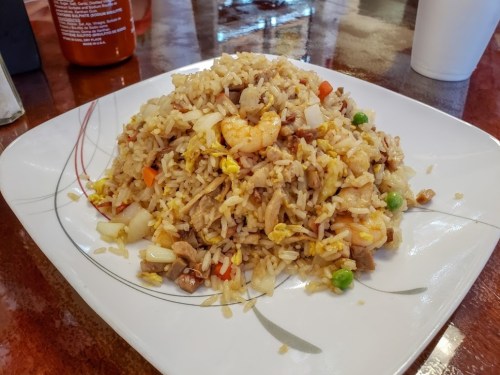 A plate of fried rice with shrimp, vegetables, and pieces of meat, garnished and served on a white dish.