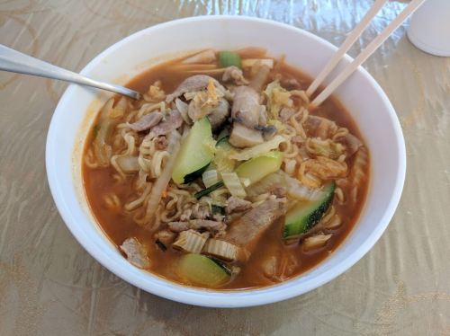 A bowl of ramen with vegetables and meat, served with chopsticks and a spoon on a textured table surface.