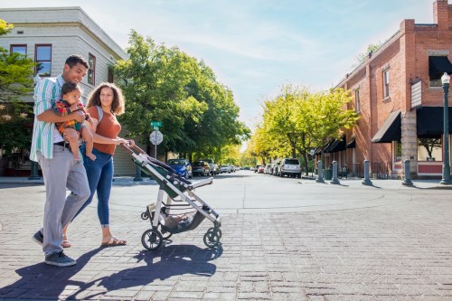 A couple walks with a baby in a stroller on a sunny street lined with trees and shops.