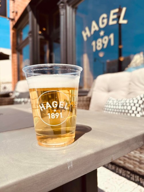 A clear plastic cup of beer sits on a table in front of a building with "HAGEL 1891" on the window.