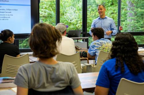 A speaker presents to an audience in a classroom with large windows and greenery outside.