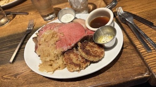 A plate of prime rib with sauerkraut, two crab cakes, and dipping sauces on a wooden table.