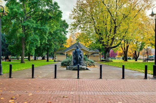 A park scene featuring a statue surrounded by trees with autumn foliage and a gazebo in the background.