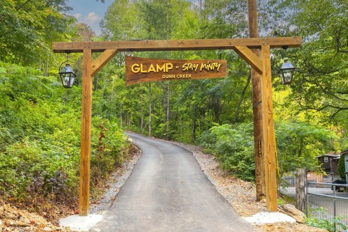 Wooden entrance sign reading "GLAMP - Say Minty" on a tree-lined road in a forested area.