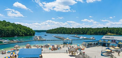 A sunny lakeside scene with boats, a sandy beach, and people enjoying the water under a blue sky with fluffy clouds.