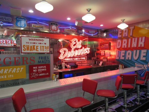 A retro diner interior with a neon sign, red bar stools, and colorful wall art promoting food and drinks.
