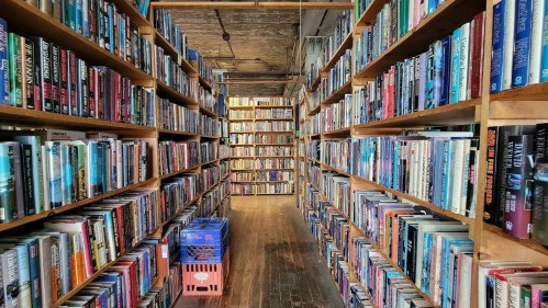 A narrow aisle between tall bookshelves filled with various books, with a crate at the end of the path.