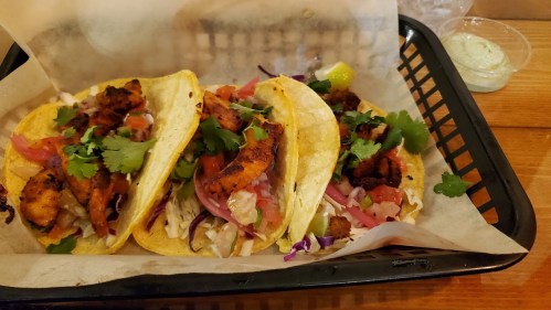 Three tacos filled with grilled chicken, cabbage, tomatoes, onions, and cilantro, served in a black basket.
