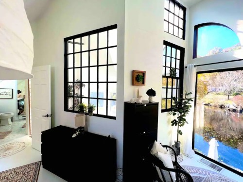 Bright interior with large windows, modern decor, and a view of a serene outdoor scene by the water.