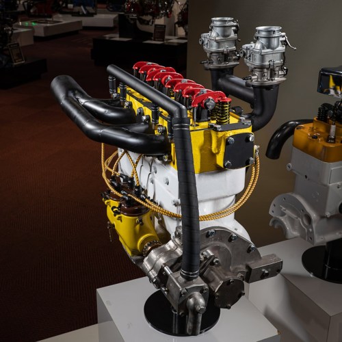 A detailed view of a colorful engine with red valve covers and multiple carburetors, displayed on a pedestal.