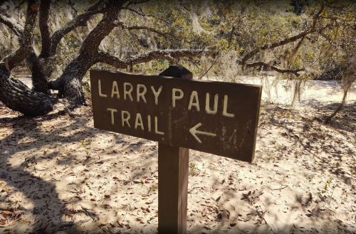 Wooden sign pointing left to "Larry Paul Trail" in a sandy, wooded area with trees and moss.