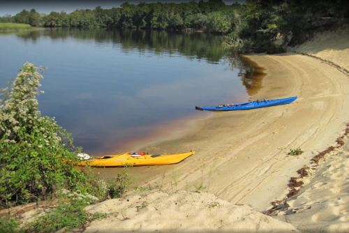 Two kayaks, one yellow and one blue, are beached on a sandy shore beside a calm river surrounded by greenery.
