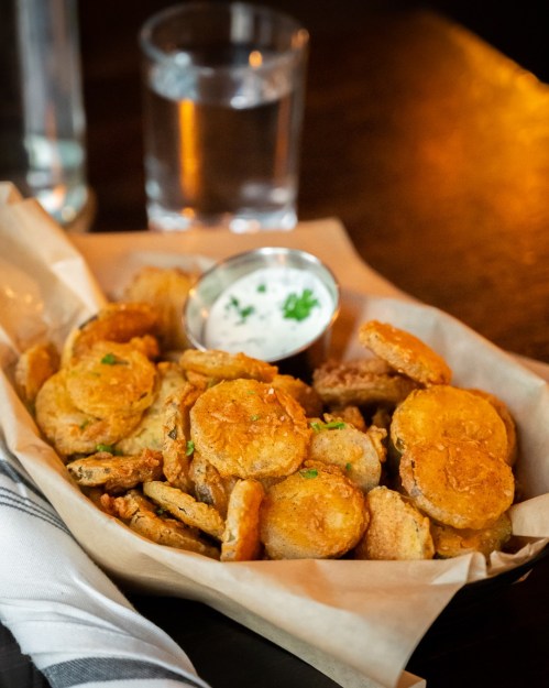 A basket of crispy fried pickles with a small bowl of ranch dressing and a glass of water in the background.