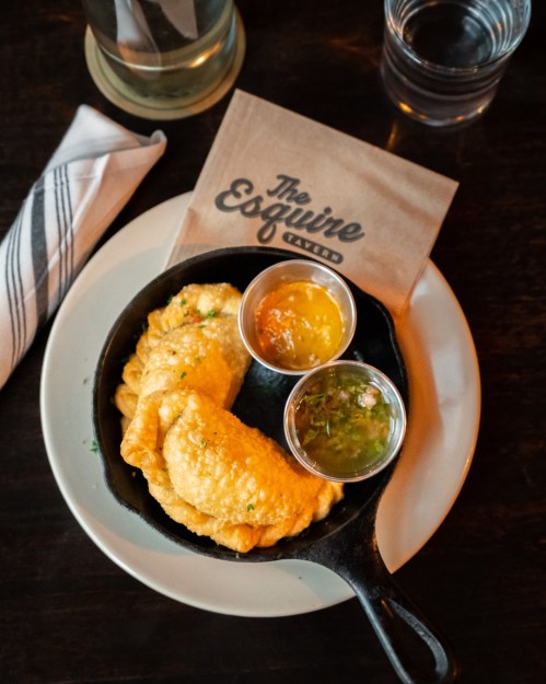 A cast iron skillet with golden fried empanadas and two dipping sauces on a plate, with a napkin and glass in the background.