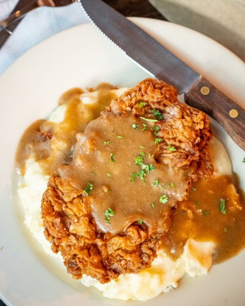A plate of fried chicken topped with gravy, served over creamy mashed potatoes, garnished with parsley.