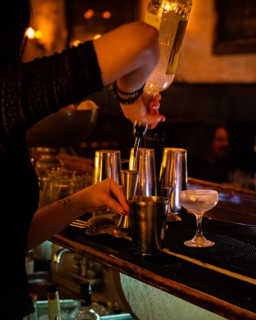 A bartender pours a drink into a shaker at a dimly lit bar, with cocktail glasses and metal shakers in the background.