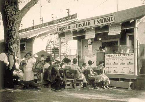 Historical photo of a eugenics and health exhibit, with people seated outside, discussing topics like birth rates and literacy.