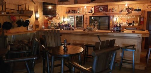 Cozy rustic bar interior with wooden furniture, shelves of bottles, and warm lighting.