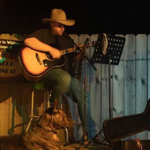 A musician in a cowboy hat plays guitar on stage, with a dog sitting nearby in a cozy outdoor setting.