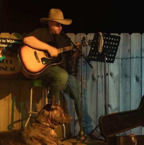 A musician in a cowboy hat plays guitar on stage, with a dog sitting nearby in a cozy outdoor setting.