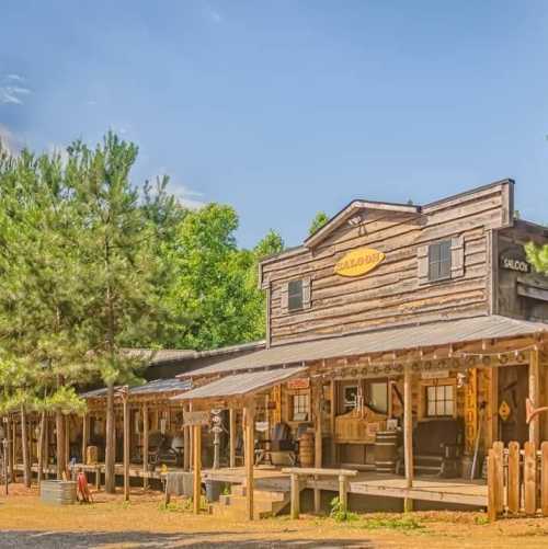 A rustic wooden saloon with a porch, surrounded by trees and a clear blue sky.