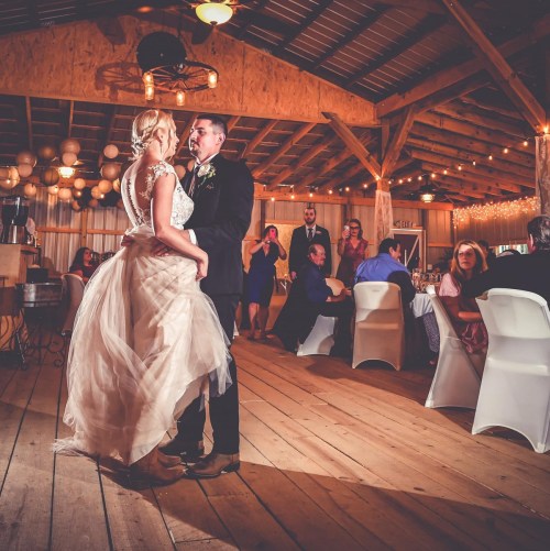 A couple dances together at their wedding reception, surrounded by guests in a rustic barn setting with warm lighting.