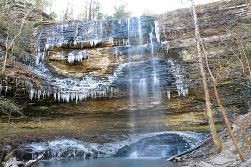 A serene waterfall cascades over rocky cliffs, surrounded by icicles and trees, reflecting in a calm pool below.