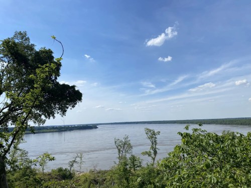 A scenic view of a river surrounded by lush greenery and a clear blue sky with scattered clouds.