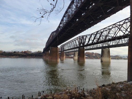 A view of two large metal bridges spanning a river, with rocky banks and a cloudy sky in the background.