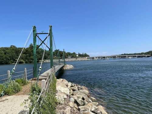 A green suspension bridge spans a calm river, with rocky banks and trees under a clear blue sky.