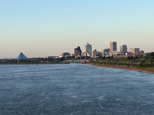 View of a city skyline along a river, featuring modern buildings and a pyramid-shaped structure in the foreground.