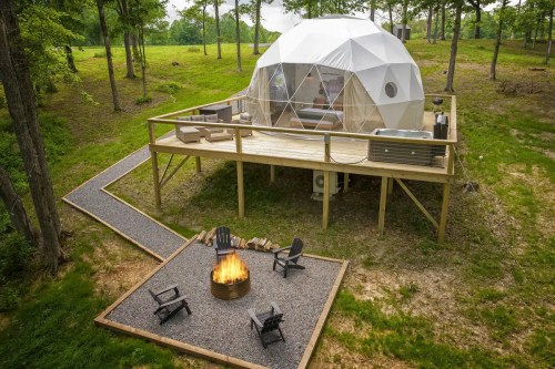A geodesic dome on a wooden deck surrounded by trees, with a fire pit and seating area in front.