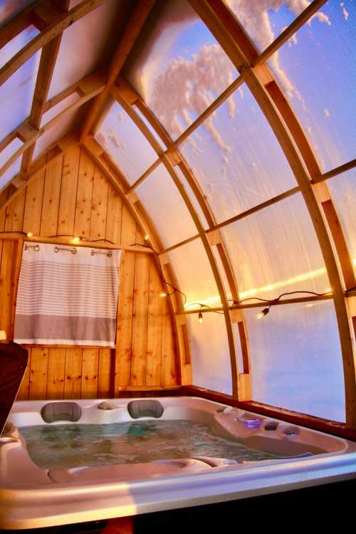 A cozy indoor hot tub surrounded by wooden walls and large windows, with soft lighting and a snowy view outside.